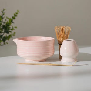 May include: A light pink ceramic matcha bowl with a spout, a bamboo whisk, a whisk holder, and a bamboo scoop. The bowl has horizontal ridges. The whisk is made of thin bamboo. The whisk holder is a small, rounded vase.