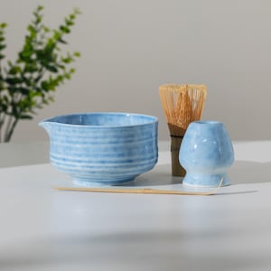 May include: A light blue ceramic matcha bowl with a spout, a bamboo whisk, a small blue ceramic stand, and a bamboo scoop. The bowl has horizontal textured lines. The items are arranged on a white surface.