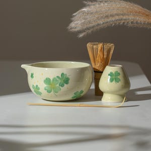 May include: A cream-colored ceramic matcha bowl with green clover designs and a spout, accompanied by a bamboo whisk, a matcha holder, and a bamboo spoon. The set is arranged on a white surface, with a neutral background.
