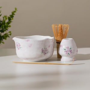 Puede incluir: Un juego de tazón, soporte y batidor de bambú para matcha de cerámica blanca. El tazón y el soporte presentan un diseño floral rosa. El batidor está hecho de bambú. El juego está diseñado para preparar té matcha.