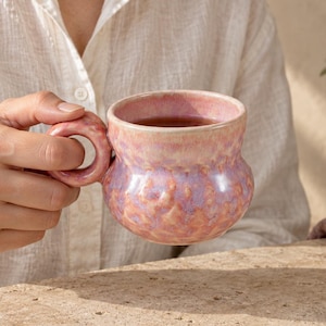 Peut inclure: Une tasse en céramique faite à la main, de forme arrondie, présentant une surface texturée avec un émail moucheté dans des tons rose, lavande et pêche. La tasse possède une anse circulaire épaisse et un corps large et bombé.