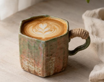 May include: A hexagonal ceramic mug with a textured, mottled green and pink glaze finish. The mug features a curved handle wrapped in thick, natural-colored jute rope and is filled with a coffee beverage topped with a white heart-shaped latte art.