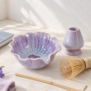 Puede incluir: Un cuenco de cerámica para matcha con borde festoneado y esmalte reactivo en tonos morado y azul, un soporte de cerámica morado a juego, un batidor de bambú y una cuchara de té de bambú. Los objetos están dispuestos sobre una superficie clara.