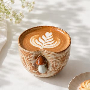 Peut inclure: Une tasse en céramique avec une finition texturée brun clair et blanc, ornée d'un petit champignon en relief sur le devant. La tasse est remplie d'un café au lait avec une mousse blanche en forme de feuille, accompagnée d'un petit biscuit.