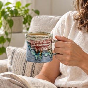Puede incluir: Una taza de cerámica con superficie texturizada que presenta una banda horizontal de esmalte rojo oscuro y blanco en la parte superior, que transiciona a un patrón de goteo azul y verde vibrante hacia la base, con asa y borde circular.