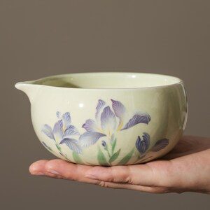 May include: A light green ceramic bowl with a pouring spout, decorated with purple and yellow iris flowers. The bowl is held in a hand, showcasing the floral design and the smooth, rounded shape.
