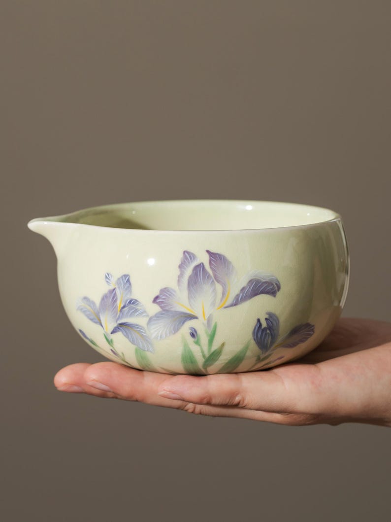 May include: A light green ceramic bowl with a pouring spout, decorated with purple and yellow iris flowers. The bowl is held in a hand, showcasing the floral design and the smooth, rounded shape.