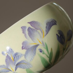 May include: A ceramic cup with a pale yellow background, adorned with hand-painted purple iris flowers with yellow centers and green leaves. The cup's design features a delicate floral pattern, showcasing a blend of artistry and nature.