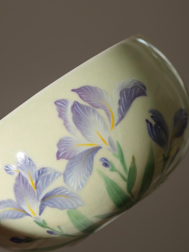 May include: A ceramic cup with a pale yellow background, adorned with hand-painted purple iris flowers with yellow centers and green leaves. The cup's design features a delicate floral pattern, showcasing a blend of artistry and nature.