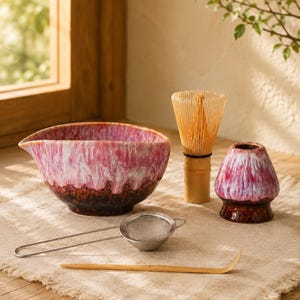 May include: A matcha tea set including a ceramic pouring bowl with a pink, white, and dark brown drip glaze, a bamboo whisk, a matching ceramic whisk holder, a stainless steel fine mesh strainer, and a curved bamboo tea scoop for preparation.