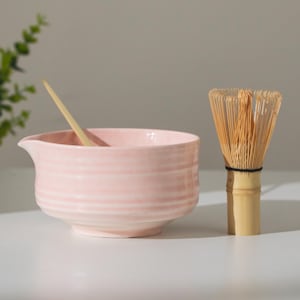 May include: A light pink ceramic matcha bowl with a wooden spoon and a bamboo whisk. The bowl has a spout and horizontal ridges. The whisk is made of thin bamboo tines bound together with a black thread.
