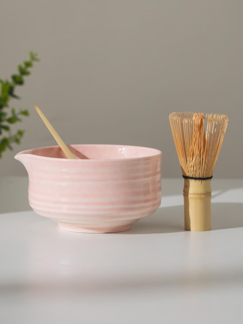 May include: A light pink ceramic matcha bowl with a wooden spoon and a bamboo whisk. The bowl has a spout and horizontal ridges. The whisk is made of thin bamboo tines bound together with a black thread.