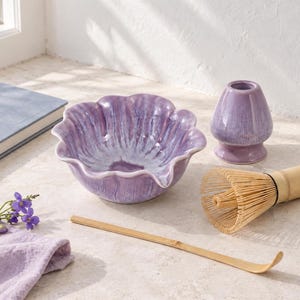 May include: A lavender ceramic matcha bowl with a scalloped edge, a matching lavender ceramic whisk holder, a light brown bamboo whisk, and a long, thin bamboo tea scoop. The set is arranged with a small sprig of purple flowers.