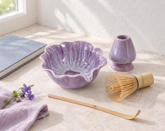 May include: A lavender ceramic matcha bowl with a scalloped edge, a matching lavender ceramic whisk holder, a light brown bamboo whisk, and a long, thin bamboo tea scoop. The set is arranged with a small sprig of purple flowers.