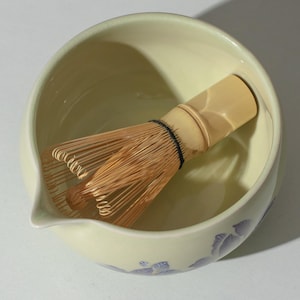 May include: A pale green ceramic matcha bowl with a spout and a bamboo whisk. The bowl has a floral design in shades of purple. The whisk is made of thin bamboo tines bound together with a black thread.