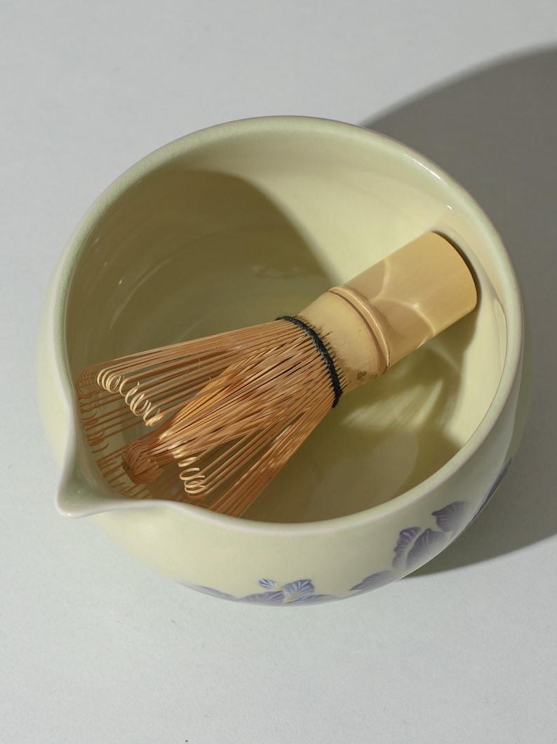 May include: A pale green ceramic matcha bowl with a spout and a bamboo whisk. The bowl has a floral design in shades of purple. The whisk is made of thin bamboo tines bound together with a black thread.