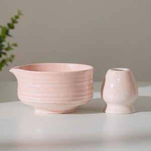 May include: A light pink ceramic bowl with a spout and a small, matching vase. The bowl has horizontal ridges, and both items are on a white surface. The items are likely for kitchen or home decor.