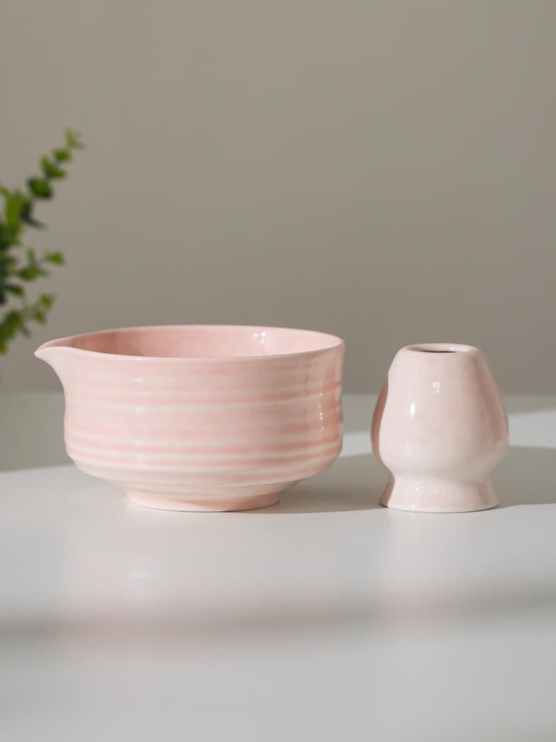 May include: A light pink ceramic bowl with a spout and a small, matching vase. The bowl has horizontal ridges, and both items are on a white surface. The items are likely for kitchen or home decor.