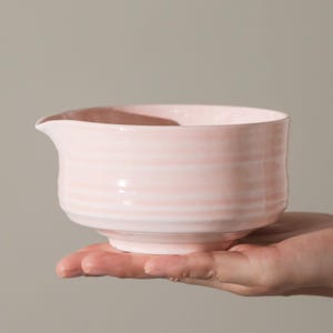 May include: A light pink ceramic bowl with a spout, held in a hand. The bowl has a ridged texture and a rounded shape. The background is a solid, neutral color.