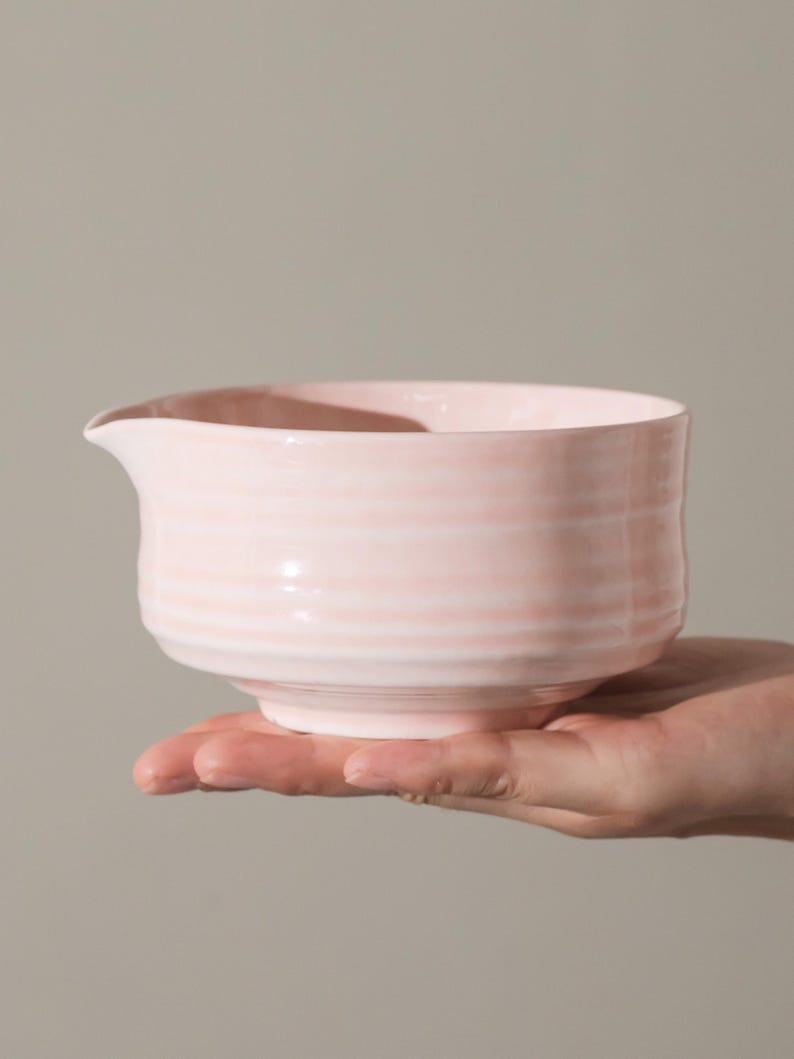 May include: A light pink ceramic bowl with a spout, held in a hand. The bowl has a ridged texture and a rounded shape. The background is a solid, neutral color.