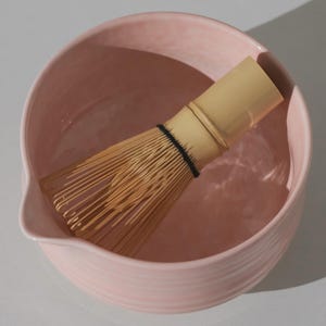 May include: A light pink ceramic matcha bowl with a bamboo whisk resting inside. The bowl has a rounded shape with a pouring spout and a ridged exterior. The whisk is made of thin bamboo tines bound together with a black band.