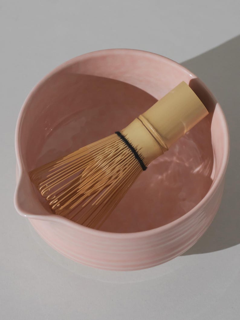 May include: A light pink ceramic matcha bowl with a bamboo whisk resting inside. The bowl has a rounded shape with a pouring spout and a ridged exterior. The whisk is made of thin bamboo tines bound together with a black band.