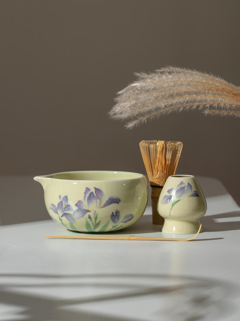 May include: A matcha tea set featuring a pale yellow ceramic bowl with a spout and purple iris floral design, a bamboo whisk, a small ceramic holder, and a bamboo scoop. The set is arranged on a white surface.