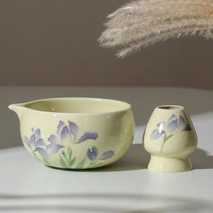 May include: A light yellow ceramic matcha bowl and whisk holder set. The bowl has a spout and is decorated with purple iris flowers and green leaves. The whisk holder has a similar design. The set is on a white surface.