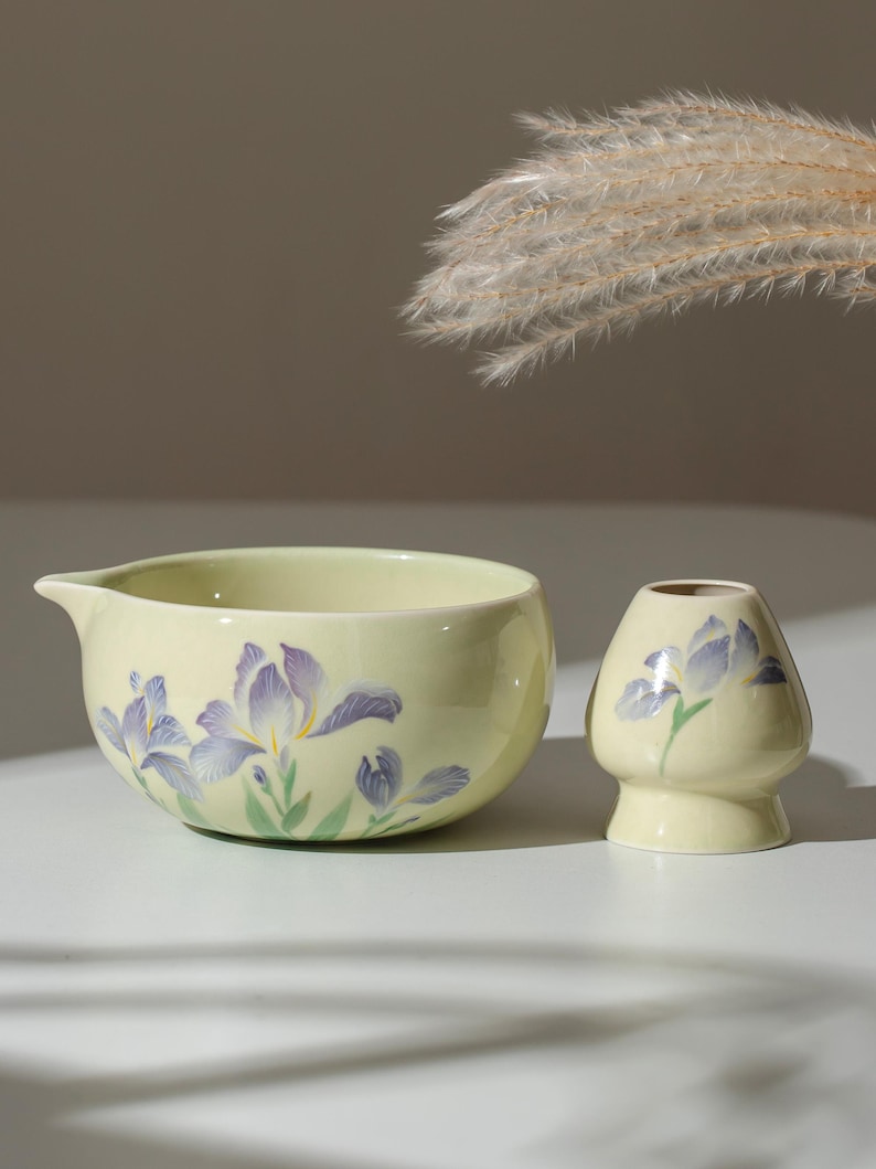 May include: A light yellow ceramic matcha bowl and whisk holder set. The bowl has a spout and is decorated with purple iris flowers and green leaves. The whisk holder has a similar design. The set is on a white surface.