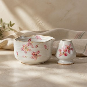 May include: A ceramic matcha bowl with a wavy rim and a matching whisk holder, both featuring a hand-painted pink cherry blossom pattern on a cream-colored glazed surface. The bowl has a wide opening and the whisk holder has a tapered shape.