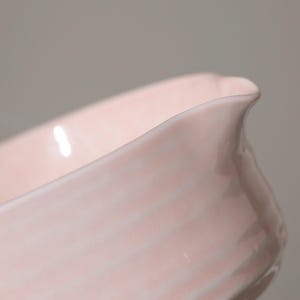 May include: A close-up of a pale pink ceramic serving dish. The dish has a curved, elongated shape with a slightly flared rim and a small spout. The surface has subtle, horizontal ridges.