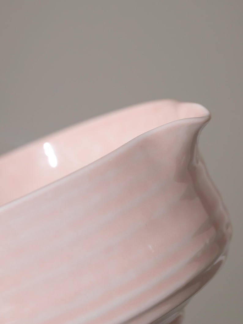 May include: A close-up of a pale pink ceramic serving dish. The dish has a curved, elongated shape with a slightly flared rim and a small spout. The surface has subtle, horizontal ridges.