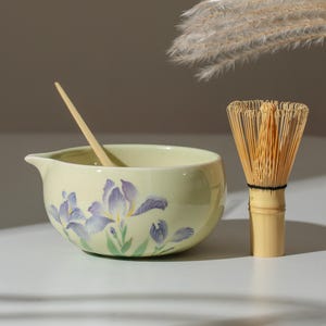 May include: A matcha tea set featuring a pale yellow ceramic bowl with a floral design of purple irises and green leaves. A bamboo whisk and a wooden spoon are also included. The bowl has a spout for easy pouring.