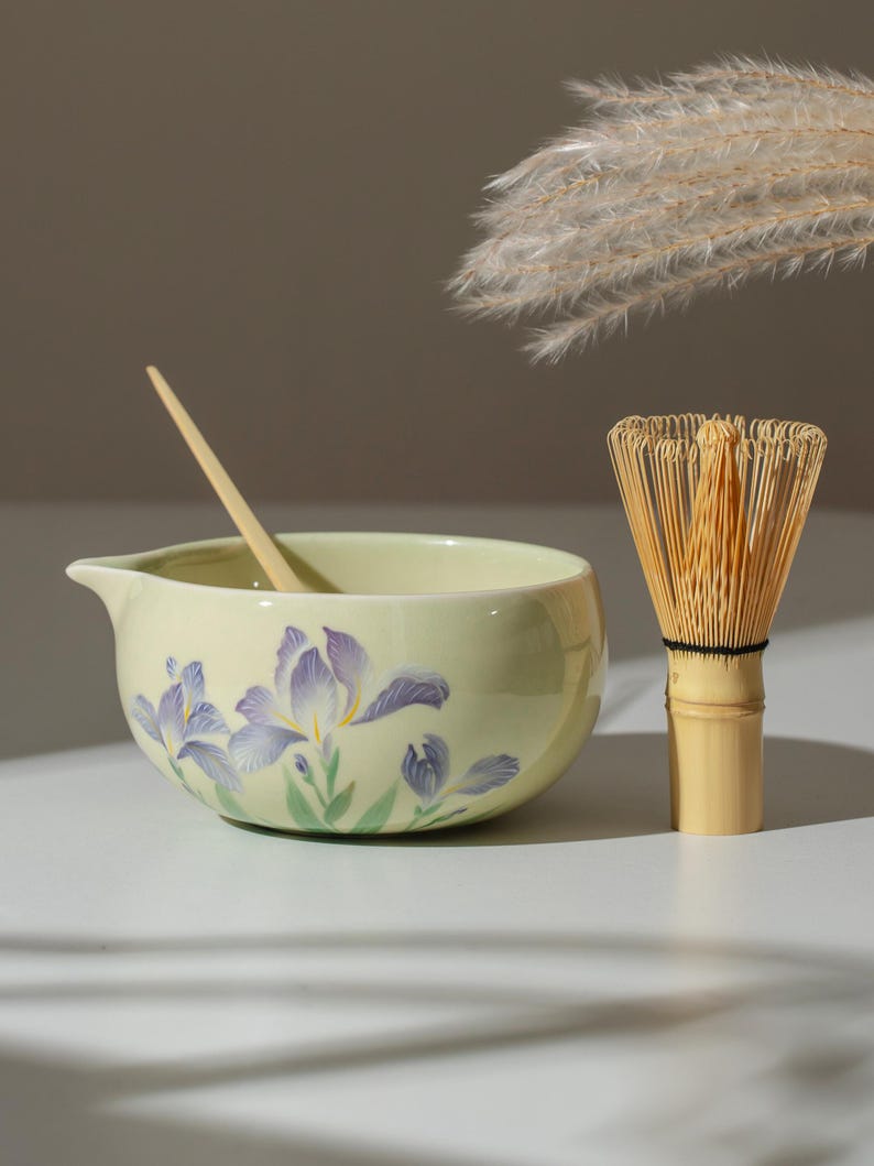 May include: A matcha tea set featuring a pale yellow ceramic bowl with a floral design of purple irises and green leaves. A bamboo whisk and a wooden spoon are also included. The bowl has a spout for easy pouring.