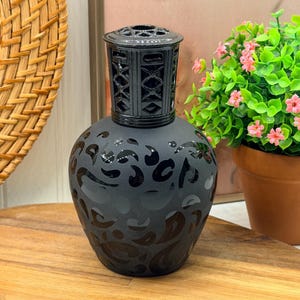 Peut inclure: Une lampe à parfum noire mate avec un corps bulbeux orné d'un motif léopard noir brillant. Le sommet de la lampe est doté d'une couronne en métal noir avec un design géométrique découpé, fixée sur le col du récipient en verre.