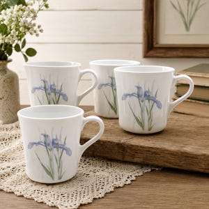 Peut inclure: Un ensemble de trois tasses en céramique blanche, chacune ornée d'une délicate illustration style aquarelle représentant une fleur d'iris bleue avec des tiges et des feuilles vertes. Les tasses ont un corps arrondi, un bord évasé et une anse.