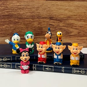 Könnte beinhalten: Eine Sammlung farbenfroher Disney-Charakterfiguren, darunter Donald Duck, Daisy Duck, Minnie Mouse, Goofy, Pluto und die drei kleinen Schweinchen. Die Figuren werden auf einer dunkelblauen Oberfläche präsentiert.