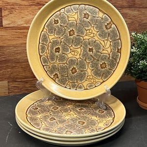 Puede incluir: Un juego de cuatro platos de cerámica amarillos con un estampado floral en tonos grises y marrones. Los platos son redondos con un borde ligeramente elevado. El diseño floral cubre el centro de los platos.