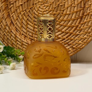 May include: An amber glass bottle with a gold-coloured metal top. The bottle has an etched design and a square base. The background features a woven wicker design.