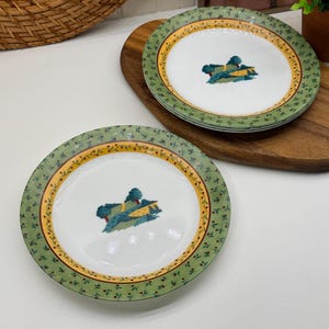 Peut inclure: Un ensemble d'assiettes rondes en céramique avec un centre blanc orné d'une petite illustration de paysage avec des arbres et des collines. Le bord présente une bande jaune et une large bordure verte à motifs de baies rouges et de feuilles.