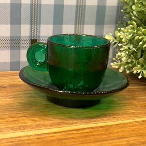 May include: A green glass teacup and saucer set. The cup has a rounded handle and the saucer has a slightly raised edge.