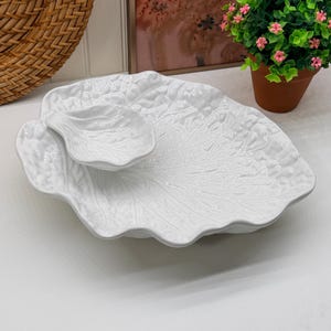 May include: A white ceramic serving dish shaped like a large cabbage leaf with a smaller, matching leaf-shaped bowl attached to the surface. The piece features a glossy finish and detailed, embossed veining throughout the entire structure.