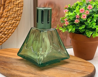 Könnte beinhalten: Eine quadratische Duftlampe aus grünem Glas mit einer facettierten, blütenblattartigen Textur an den Seiten. Die Lampe verfügt über eine metallisch grüne, belüftete Kappe an der Oberseite und einen weißen Docht im Inneren des smaragdgrünen Glaskörpers.