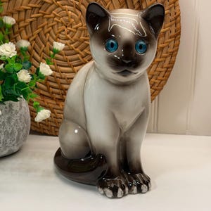 Peut inclure: Figurine de chat siamois en céramique avec des yeux bleus vifs. Le chat est représenté en position assise, avec une finition brillante et des détails soignés. La palette de couleurs comprend des nuances de marron, de crème et de noir. Objet décoratif.