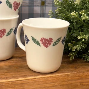 Peut inclure: Une tasse en céramique blanche avec un motif floral vert, rouge et bleu. La tasse a une anse et est posée sur une surface en bois.