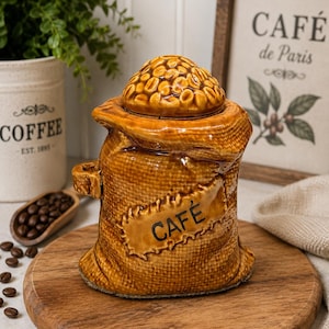 Peut inclure: Un pot en céramique marron en forme de sac de café en toile de jute avec un couvercle texturé représentant un tas de grains de café. L'avant présente un écusson rectangulaire avec le mot CAFÉ imprimé en noir. Le pot possède une petite anse.