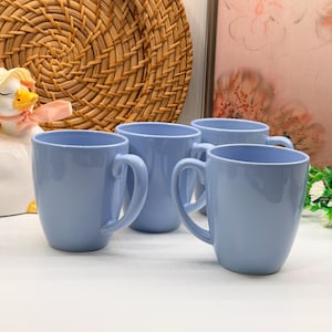 Peut inclure: Un ensemble de quatre tasses en céramique bleu clair avec une finition lisse et brillante et des anses incurvées. Chaque tasse présente une silhouette simple et arrondie avec une couleur bleu pâle uniforme sur toutes les surfaces.