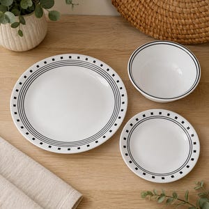 Puede incluir: Un juego de vajilla blanco que consta de un plato grande, un plato pequeño y un cuenco. Cada pieza presenta un diseño de borde negro compuesto por líneas circulares concéntricas y un patrón repetitivo de pequeños cuadrados negros.