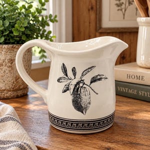May include: A cream-colored ceramic pitcher featuring a black and white botanical illustration of a lemon with leaves on the front. The base of the pitcher is decorated with a detailed black patterned border consisting of small repeating leaf motifs.