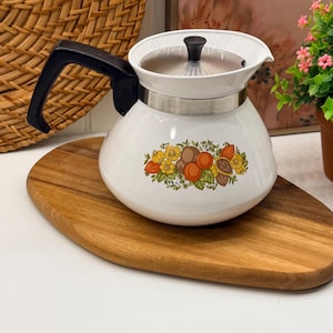 Puede incluir: Una tetera blanca con asa negra y tapa de metal plateado con pomo negro. El cuerpo de la tetera está decorado en la parte frontal con una colorida ilustración botánica de frutas naranjas, amarillas y marrones junto a hojas de color verde.